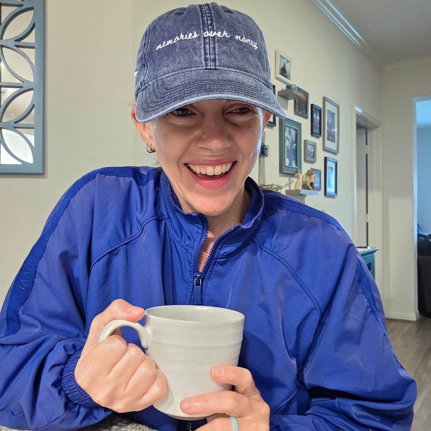 Person wearing a blue jacket and travel hat with the words "memories over money" on it, holding a mug indoors.