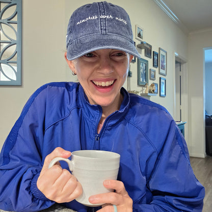 Person wearing a blue jacket and travel hat with the words "memories over money" on it, holding a mug indoors.