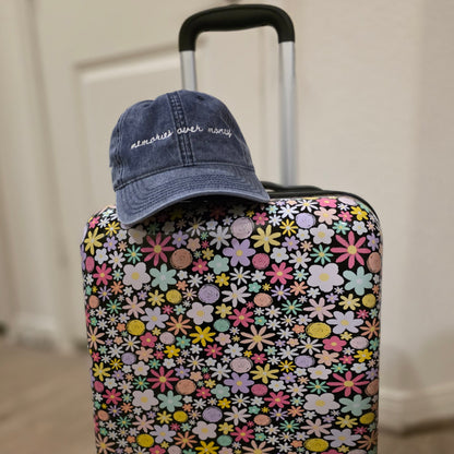 Floral-patterned suitcase with a blue travel themed hat with words "memories over money" on top, against a neutral background
