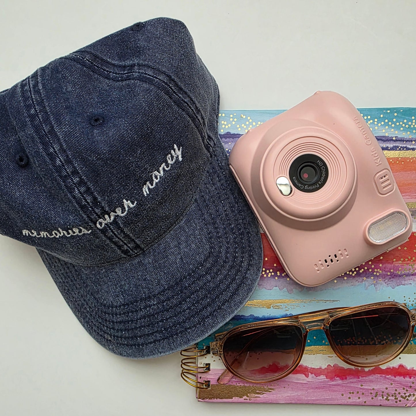 Navy cap with 'memories over money' text, pink camera, sunglasses, and colorful book on a white surface
