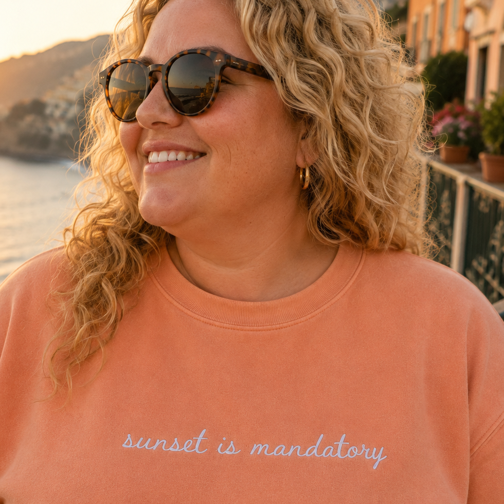 A smiling woman with curly blonde hair and sunglasses wearing a peach travel themed crewneck sweatshirt with the text 'sunset is mandatory' embroidered in white script, set against a coastal background at sunset.
