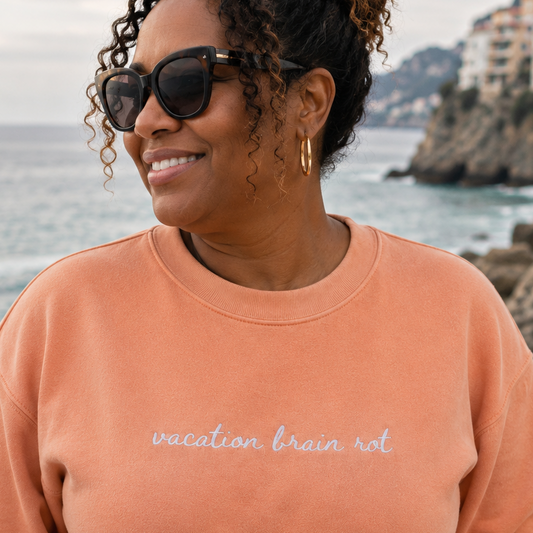 A woman wearing a peach crewneck sweatshirt with the text 'vacation brain rot' embroidered in white script, standing outdoors by the sea with a coastal town in the background.