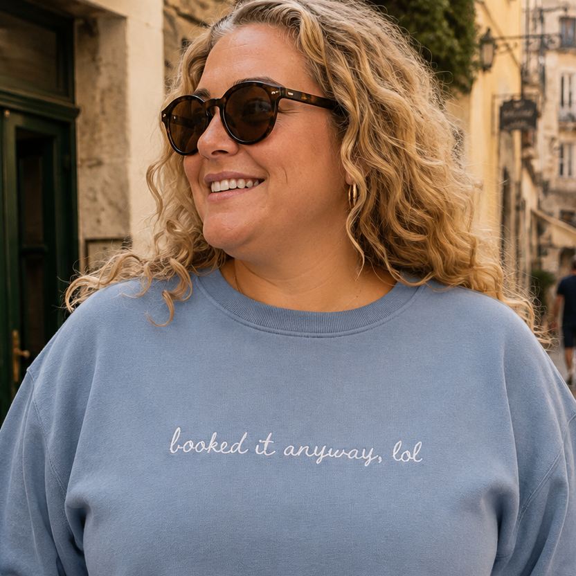 A woman wearing a dusty blue travel themed crewneck sweatshirt with the phrase 'booked it anyway, lol' embroidered in white script across the chest, standing in a cobblestone street.