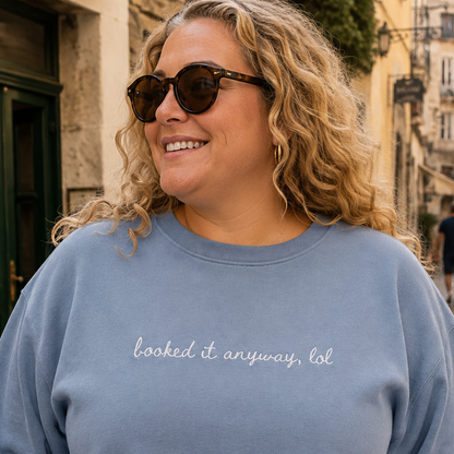 A woman wearing a dusty blue travel themed crewneck sweatshirt with the phrase 'booked it anyway, lol' embroidered in white script across the chest, standing in a cobblestone street.