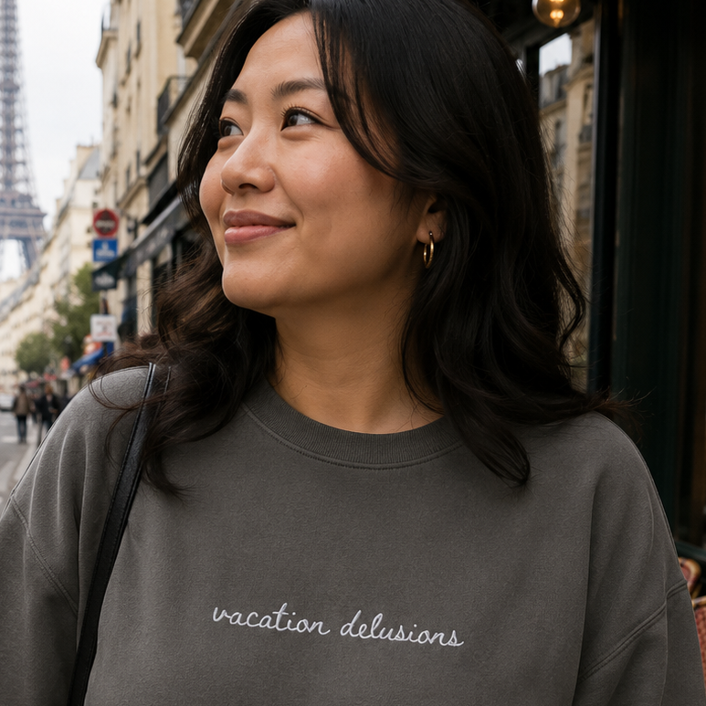 A woman wearing a dark gray crewneck sweatshirt with 'vacation delusions' embroidered in white script across the chest, standing on a Parisian street with the Eiffel Tower in the background.