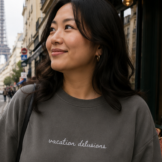 A woman wearing a dark gray crewneck sweatshirt with 'vacation delusions' embroidered in white script across the chest, standing on a Parisian street with the Eiffel Tower in the background.