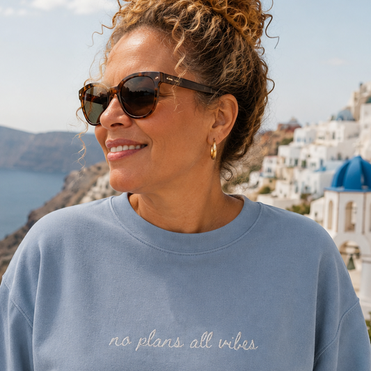 A woman wearing a light blue crewneck sweatshirt with 'no plans all vibes' embroidered in white script across the chest, posing outdoors with a scenic view of Santorini in the background.