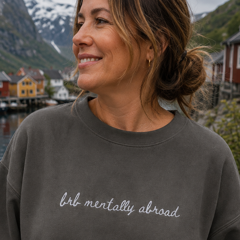 A woman wearing a dark gray crewneck sweatshirt with the phrase 'brb mentally abroad' written in white script across the chest, posing outdoors with a scenic backdrop of mountains, water, and red houses.