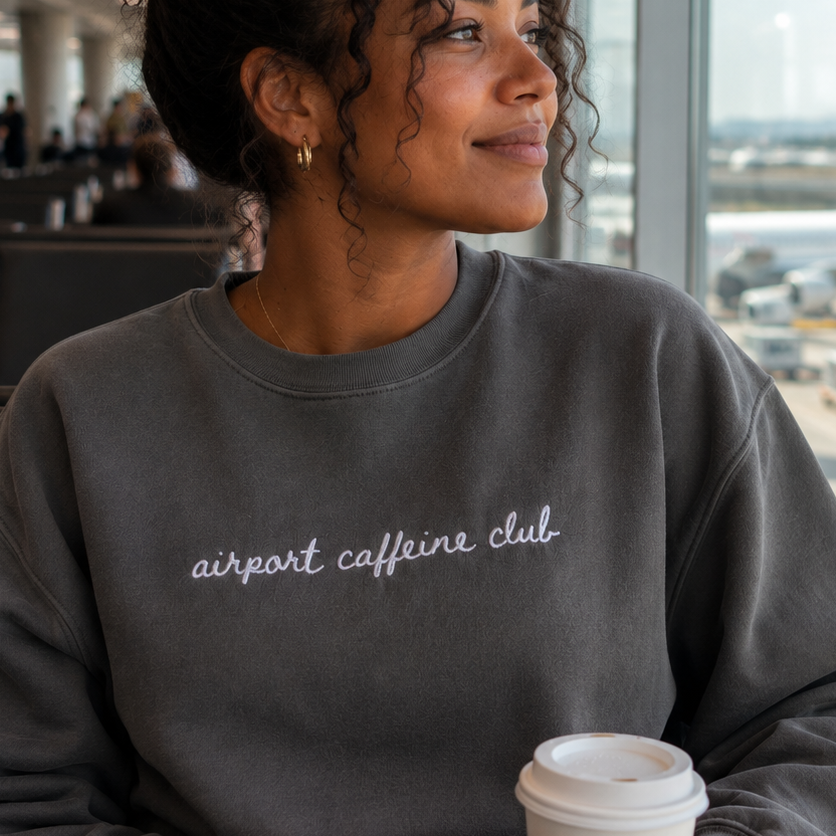 A woman sitting in an airport lounge holding a coffee cup, wearing a dark gray sweatshirt with 'airport caffeine club' embroidered in white script across the chest.