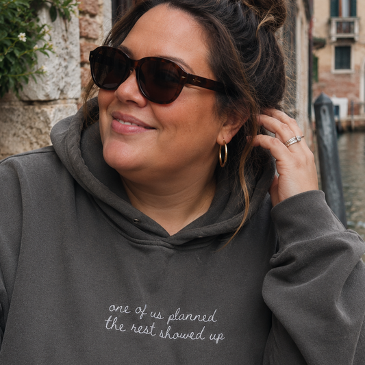 A woman wearing a dark gray hoodie with the phrase 'one of us planned the rest showed up' in white script, photographed outdoors near a canal with stone buildings and plants in the background.