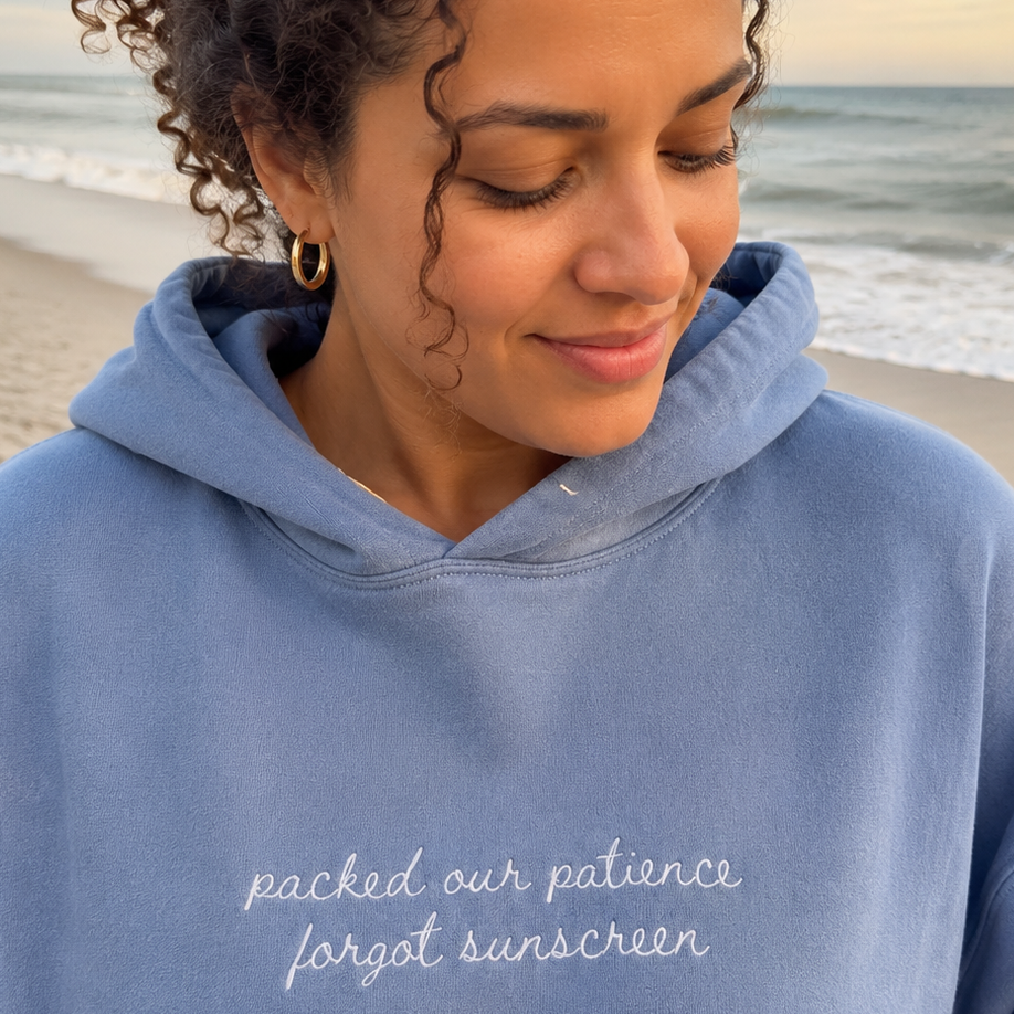 A woman wearing a dusty blue hoodie with white embroidered text reading 'packed our patience forgot sunscreen' stands on a beach with the ocean and sky in the background.