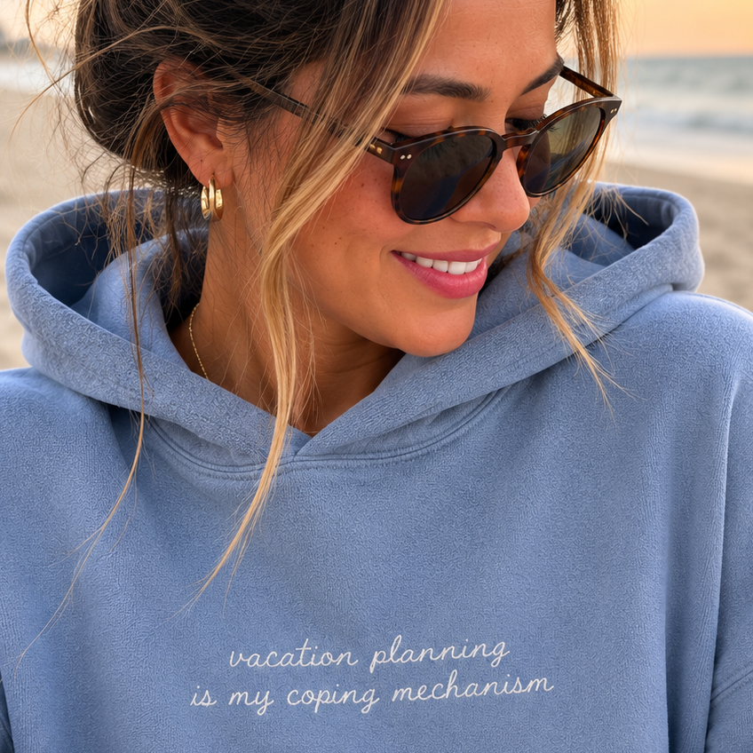 A woman wearing a dusty blue hoodie with the text 'vacation planning is my coping mechanism' in white script, sitting on a beach at sunset.