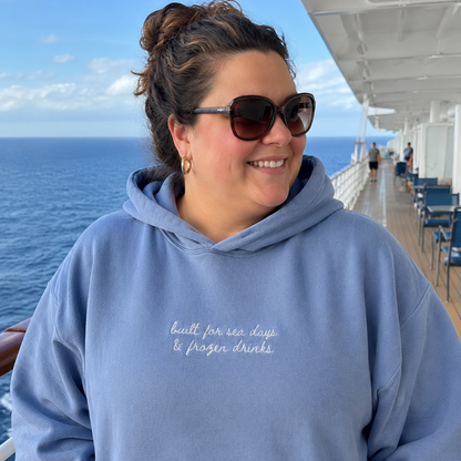 A woman wearing a light blue hoodie with white cursive text reading 'built for sea days & frozen drinks' stands on a cruise ship deck with the ocean in the background.