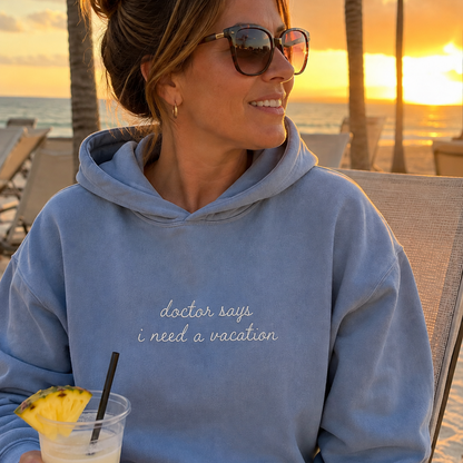 A woman wearing a light blue hoodie with the text 'doctor says i need a vacation' in white script, sitting on a beach chair at sunset with palm trees and ocean in the background.