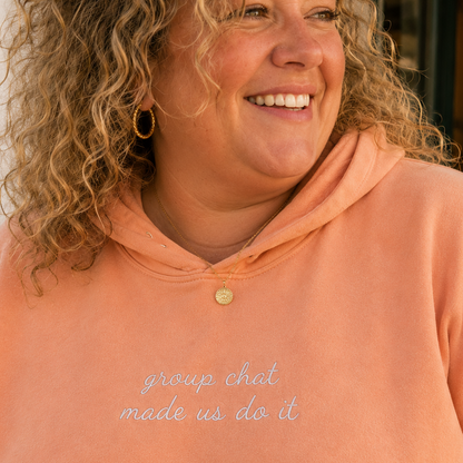 A smiling woman with curly blonde hair wearing a peach-colored hoodie with the phrase 'group chat made us do it' embroidered in light blue script on the front.
