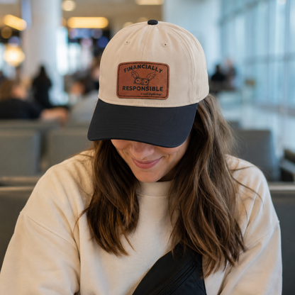 A woman wearing a cream and navy baseball cap with a brown leather patch that reads 'FINANCALLY RESPONSIBLE (until flights drop)'