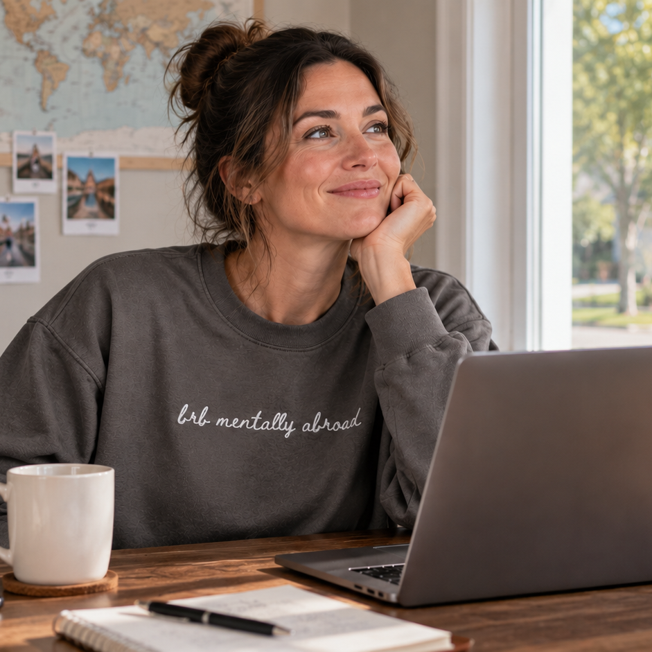 A woman with her hair in a bun sits at a wooden desk in a bright home office, wearing a dark gray sweatshirt with the white script text 'brb mentally abroad'. She has a laptop open in front of her, with a white mug, notebook, and pens on the desk.
