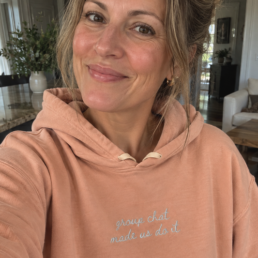A woman wearing a dusty peach-pink hoodie with the phrase 'group chat made us do it' embroidered in light blue on the chest, taking a selfie in a bright living room.