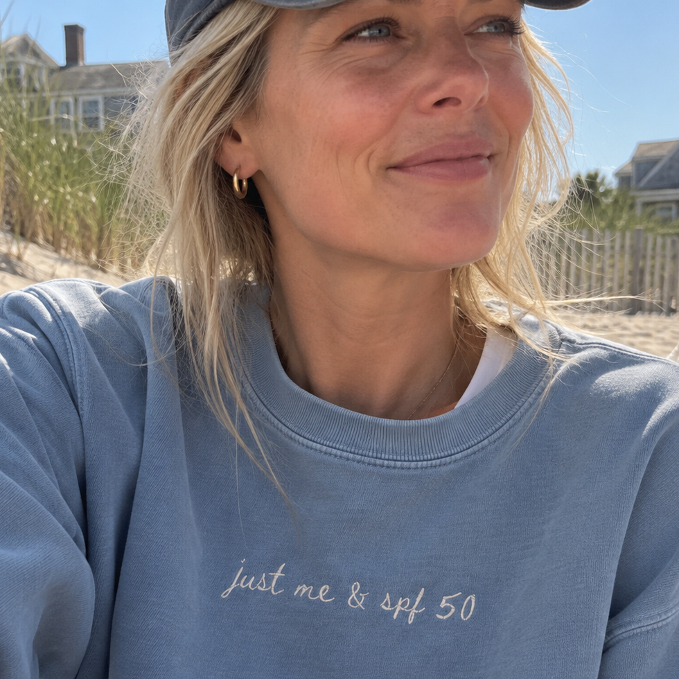 A woman wearing a light blue crewneck sweatshirt with the text 'just me & spf 50' embroidered on the chest, photographed outdoors on a beach with dunes and houses in the background.
