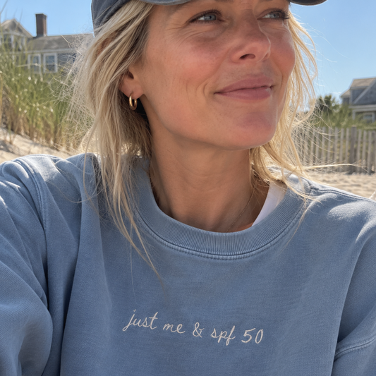 A woman wearing a light blue crewneck sweatshirt with the text 'just me & spf 50' embroidered on the chest, photographed outdoors on a beach with dunes and houses in the background.
