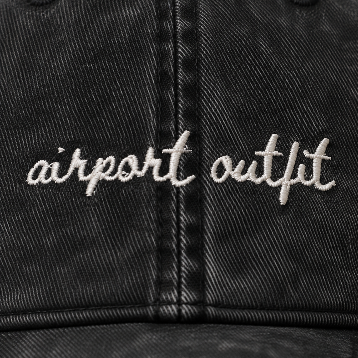 Close-up of a black washed baseball cap with 'airport outfit' embroidered in white script across the front panels.