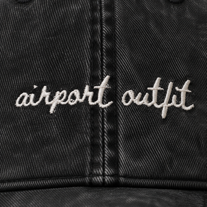 Close-up of a black washed baseball cap with 'airport outfit' embroidered in white script across the front panels.
