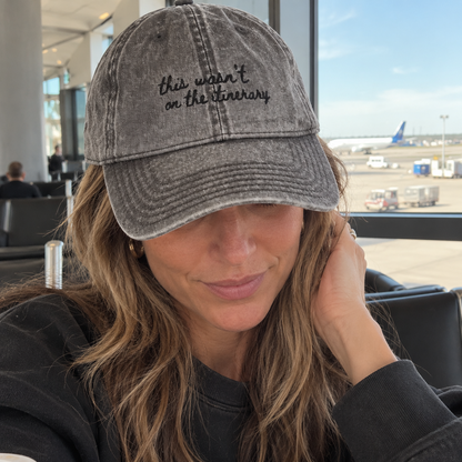 A woman wearing a gray washed baseball cap with the embroidered text 'this wasn't on the textbook' in black script, photographed in a bright airport or cafe setting with a coffee cup in the foreground.