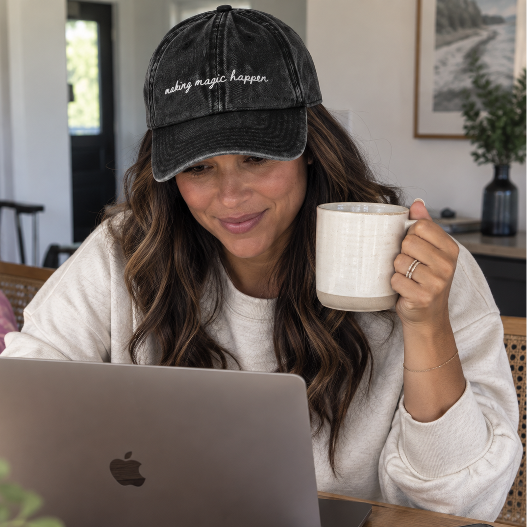 A woman sitting at a wooden table wearing a black velvet baseball cap with the embroidered text 'making magic happen.' She is holding a white ceramic mug and looking at a laptop computer.
