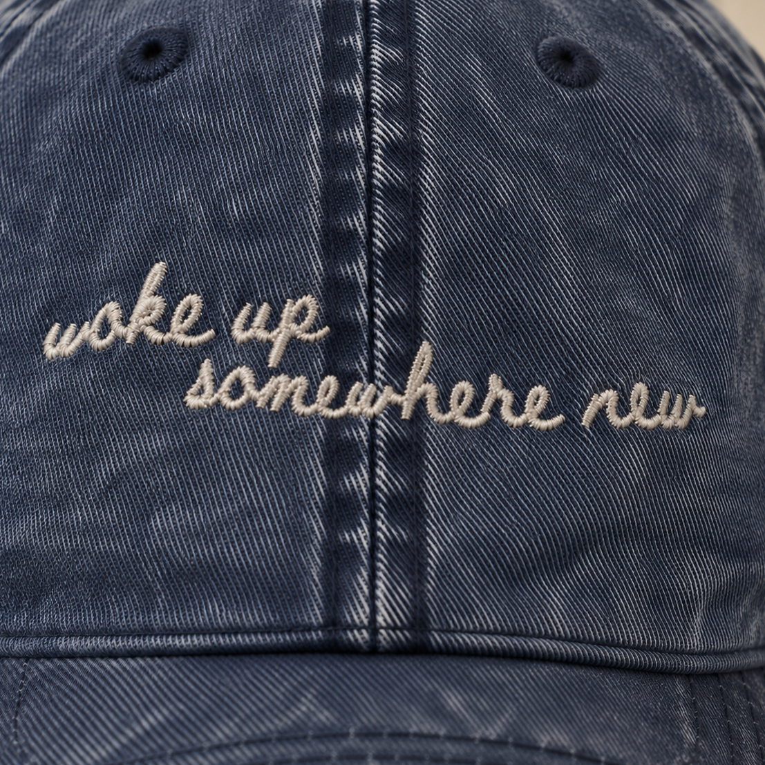 A close-up of a washed navy blue denim baseball cap featuring the phrase 'woke up somewhere new' embroidered in beige script across the front.
