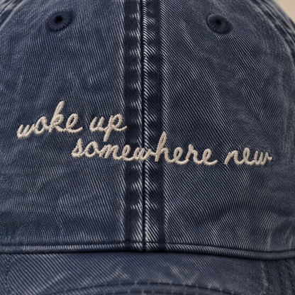 A close-up of a washed navy blue denim baseball cap featuring the phrase 'woke up somewhere new' embroidered in beige script across the front.