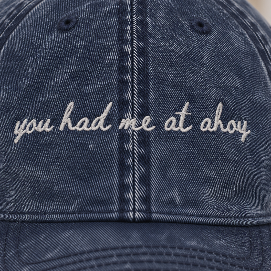 A close-up of a dark blue acid-wash cotton baseball cap with white embroidered text reading 'you had me at ohoy' on the front panel.