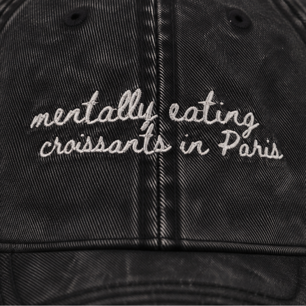 A close-up of a black washed cotton baseball cap featuring white embroidered text that reads 'mentally eating croissants in Paris'.