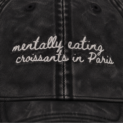 A close-up of a black washed cotton baseball cap featuring white embroidered text that reads 'mentally eating croissants in Paris'.