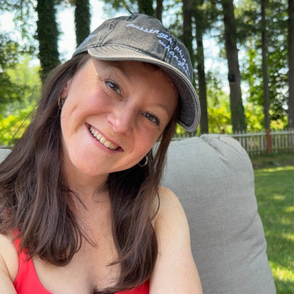 A smiling woman wearing a gray baseball cap and a red tank top, seated outdoors on a gray cushion with trees and grass in the background.