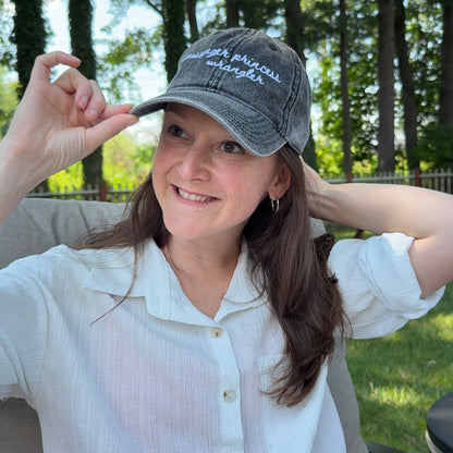 A woman wearing a gray washed baseball cap with white embroidered text reading 'Breathe Easy Whippet' sits outdoors in a garden setting, adjusting the brim of the hat.
